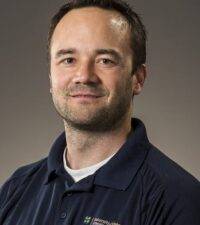 white man smiling slightly in a blue sweater and white t-shirt amid a brown backdrop