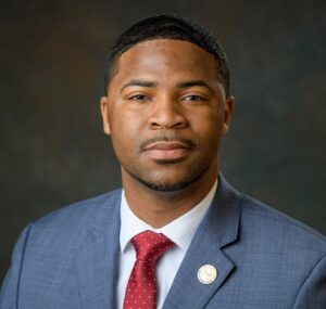 headshot of a black man in a light blue blazer, white collared shirt and red tie