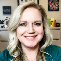 white woman with long blonde hair in a green sweater with an olive scarf smiling in her office