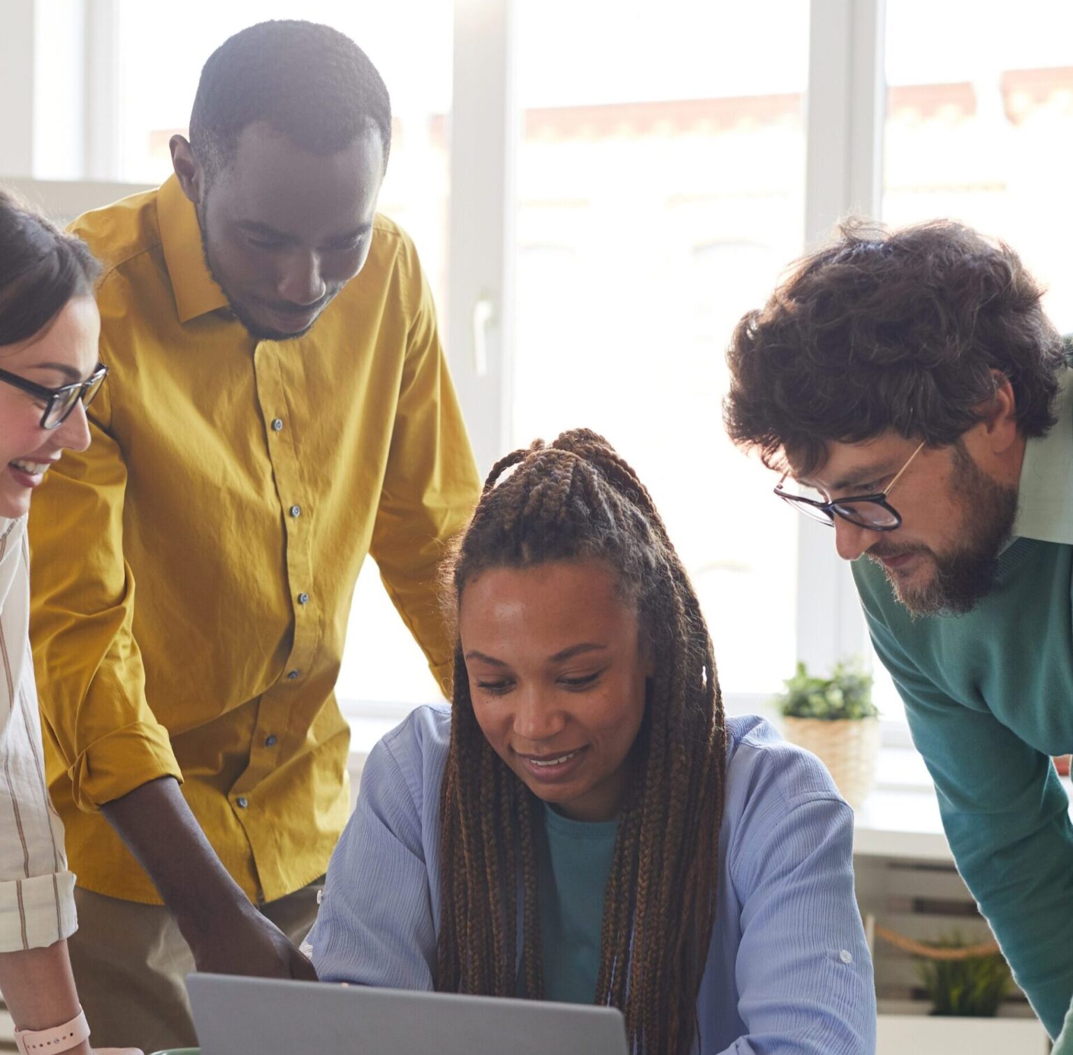A diverse group of five people collaborating around a laptop in a bright, modern office space. The group appears engaged, with one person typing while others observe and contribute. The table is cluttered with notebooks, pens, and coffee cups, emphasizing a casual yet productive work environment.
