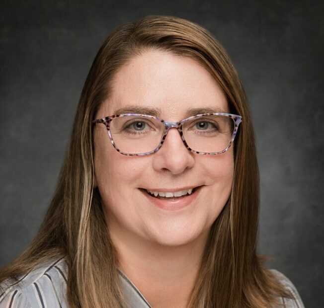 white woman with long light brown hair wearing glasses and a grey sweater smiling