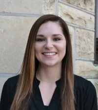 white woman smiling with long brown hair and a black shirt standing in front of a grey brick building