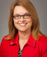 white woman with long red hair smiling wearing glasses and a red shirt amid a grey backdrop