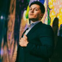 Headshot of Miguel Quinones, an award-winning presenter, looking confident in from of a multi-colored background wall wearing a professional suit.