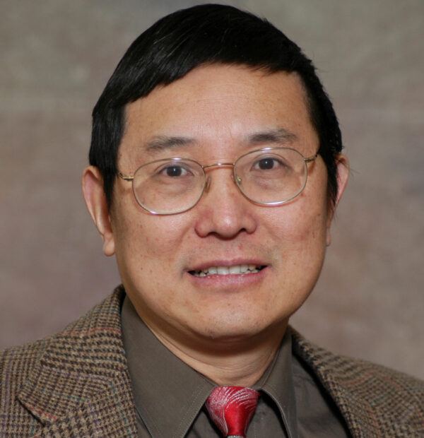 Asian man smiling wearing glasses and a brown suit with a dark collared shirt and red tie
